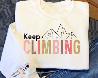 Puede incluir: Sudadera blanca con el texto "Keep Climbing" en letras rosa, marrón y doradas con un gráfico de cordillera de montañas sobre el texto. La manga izquierda tiene el texto "You can do hard things" en negro con un pequeño corazón.