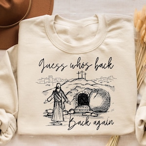 May include: A beige sweatshirt with a black graphic print of a person walking out of a tomb. The text "Guess who's back back again" is printed below the graphic.