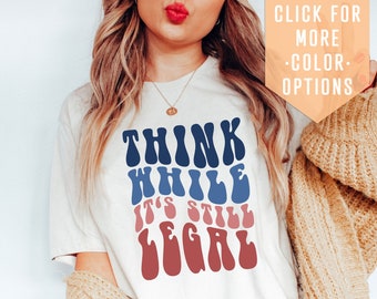 May include: White t-shirt with a blue, red, and pink graphic that says "Think While It's Still Legal" in a retro font. The shirt is being modeled by a person wearing jeans and a cardigan.