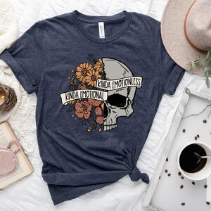 Pu&ograve; includere: Una t-shirt blu navy con un motivo a forma di teschio. Met&agrave; del teschio &egrave; ricoperta di fiori e il testo "Kinda Emotional", mentre l'altra met&agrave; &egrave; liscia con il testo "Kinda Emotionless".