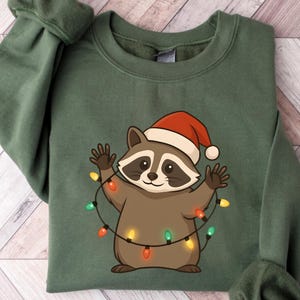 Puede incluir: Una sudadera verde con un mapache de dibujos animados que lleva un gorro de Pap&aacute; Noel y est&aacute; envuelto en luces navide&ntilde;as. El mapache es marr&oacute;n y canela con una m&aacute;scara negra. Las luces son rojas, verdes y amarillas. La sudadera est&aacute; doblada.
