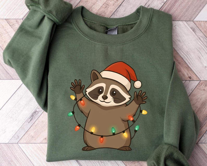 Puede incluir: Una sudadera verde con un mapache de dibujos animados que lleva un gorro de Pap&aacute; Noel y est&aacute; envuelto en luces navide&ntilde;as. El mapache es marr&oacute;n y canela con una m&aacute;scara negra. Las luces son rojas, verdes y amarillas. La sudadera est&aacute; doblada.