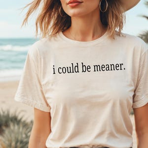 May include: Cream-colored t-shirt with the phrase "i could be meaner" printed in black. The shirt has a crew neck and short sleeves. The background shows a beach scene with the ocean and palm trees.