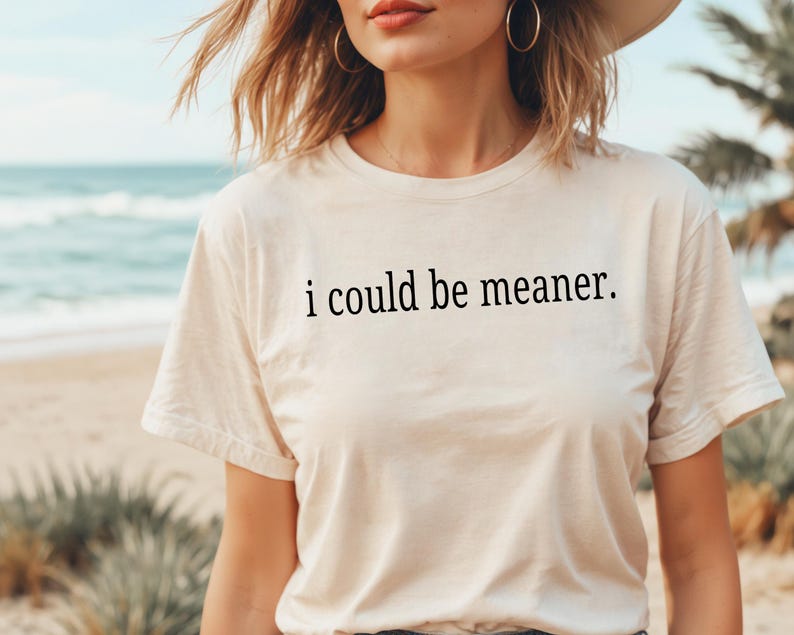 May include: Cream-colored t-shirt with the phrase "i could be meaner" printed in black. The shirt has a crew neck and short sleeves. The background shows a beach scene with the ocean and palm trees.