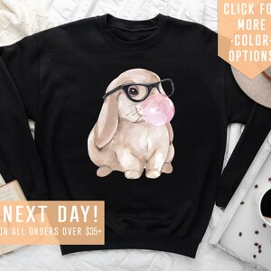 May include: Black sweatshirt with a watercolor illustration of a brown bunny wearing glasses and blowing a pink bubble. The text "SHIPS NEXT DAY! FREE SHIPPING ON ALL ORDERS OVER $35+" is at the bottom of the image.