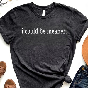 May include: Dark gray t-shirt with the phrase "i could be meaner" in white lettering. The shirt is styled with rolled-up sleeves and a knotted side. Other items in the image include a brown hat and blue jeans.