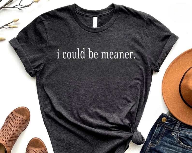 May include: Dark gray t-shirt with the phrase "i could be meaner" in white lettering. The shirt is styled with rolled-up sleeves and a knotted side. Other items in the image include a brown hat and blue jeans.