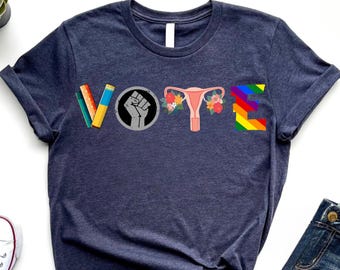 Può includere: Una maglietta blu navy con la parola "VOTE" scritta usando diversi elementi grafici, tra cui matite, un pugno, un utero e un arcobaleno.