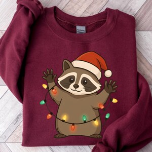 Puede incluir: Sudadera de color burdeos con un mapache de dibujos animados que lleva un gorro de Pap&aacute; Noel y sostiene una cadena de luces navide&ntilde;as de colores. El mapache es marr&oacute;n y canela con una expresi&oacute;n sonriente. La sudadera es de material suave.