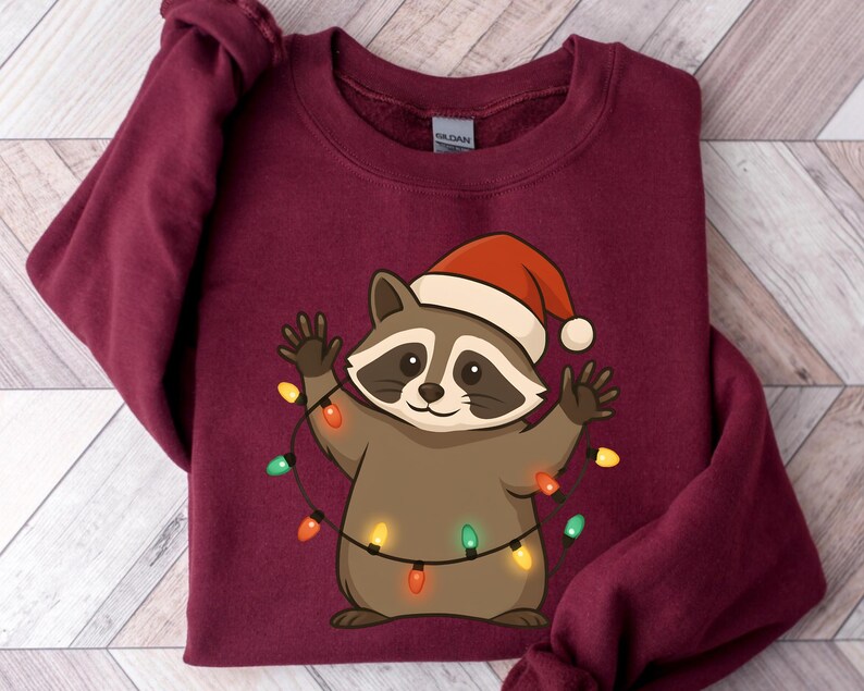 Puede incluir: Sudadera de color burdeos con un mapache de dibujos animados que lleva un gorro de Pap&aacute; Noel y sostiene una cadena de luces navide&ntilde;as de colores. El mapache es marr&oacute;n y canela con una expresi&oacute;n sonriente. La sudadera es de material suave.