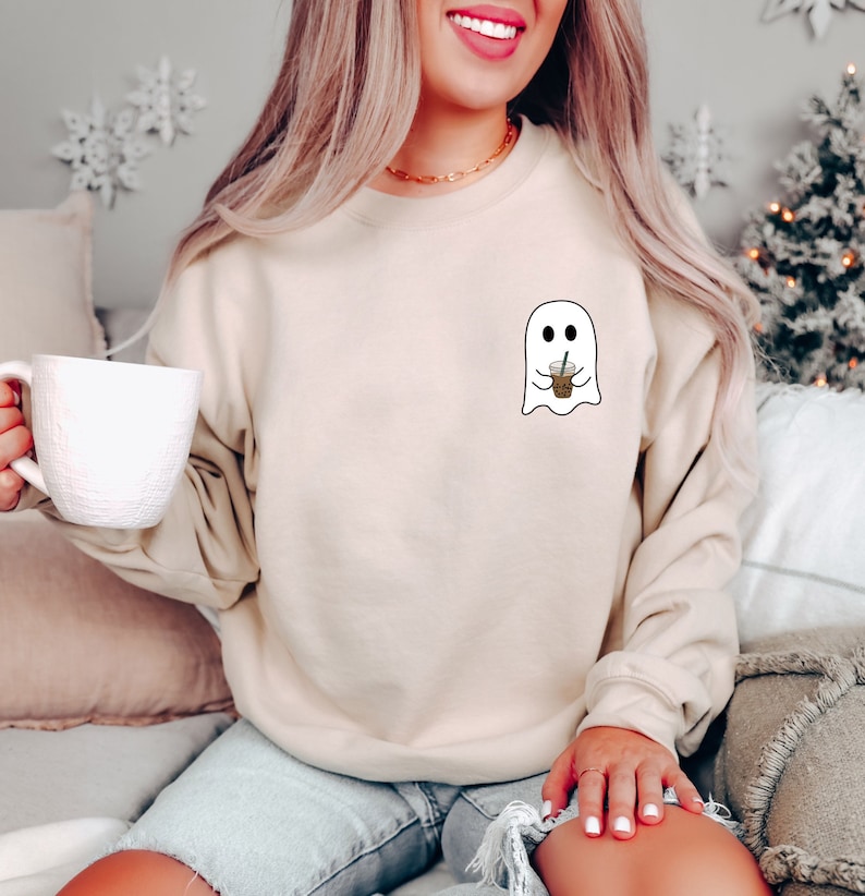 Puede incluir: Sudadera beige con un peque&ntilde;o dise&ntilde;o de fantasma blanco sosteniendo una taza de caf&eacute;. El fantasma tiene ojos negros y est&aacute; delineado en negro. La sudadera tiene mangas largas y cuello redondo. En la imagen se sostiene una taza blanca.