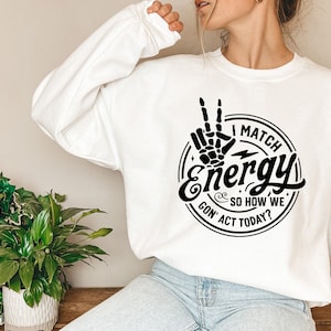 Op de afbeelding: Wit sweatshirt met een zwarte grafische print van een hand die een peace-teken maakt met de tekst "I Match Energy & So How We Gon' Act Today?"