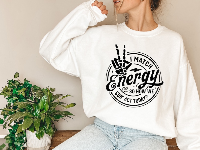 Op de afbeelding: Wit sweatshirt met een zwarte grafische print van een hand die een peace-teken maakt met de tekst "I Match Energy & So How We Gon' Act Today?"