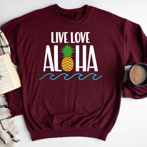 May include: Burgundy sweatshirt with the words "LIVE. LOVE. ALOHA" in white, a yellow pineapple, and blue waves. A book, glasses, and a mug of coffee are next to the sweatshirt.