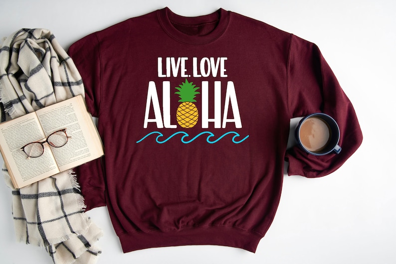 May include: Burgundy sweatshirt with the words "LIVE. LOVE. ALOHA" in white, a yellow pineapple, and blue waves. A book, glasses, and a mug of coffee are next to the sweatshirt.