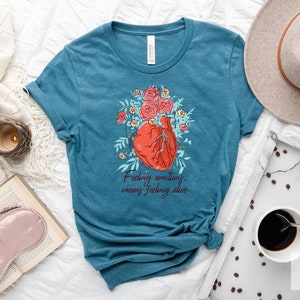 May include: Teal blue t-shirt with a graphic of a heart with flowers and the text "Feeling emotions means feeling alive."
