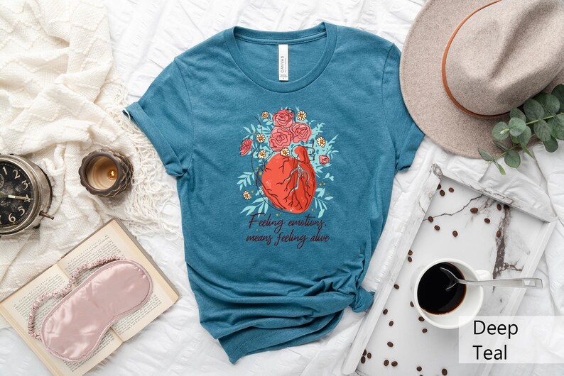 May include: Teal blue t-shirt with a graphic of a heart with flowers and the text "Feeling emotions means feeling alive."