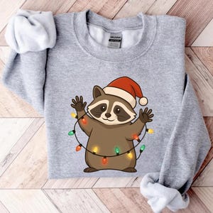 Puede incluir: Sudadera gris claro con un mapache de dibujos animados con un gorro de Pap&aacute; Noel y una guirnalda de luces navide&ntilde;as de colores. El mapache tiene un cuerpo marr&oacute;n, una m&aacute;scara negra y est&aacute; sonriendo.