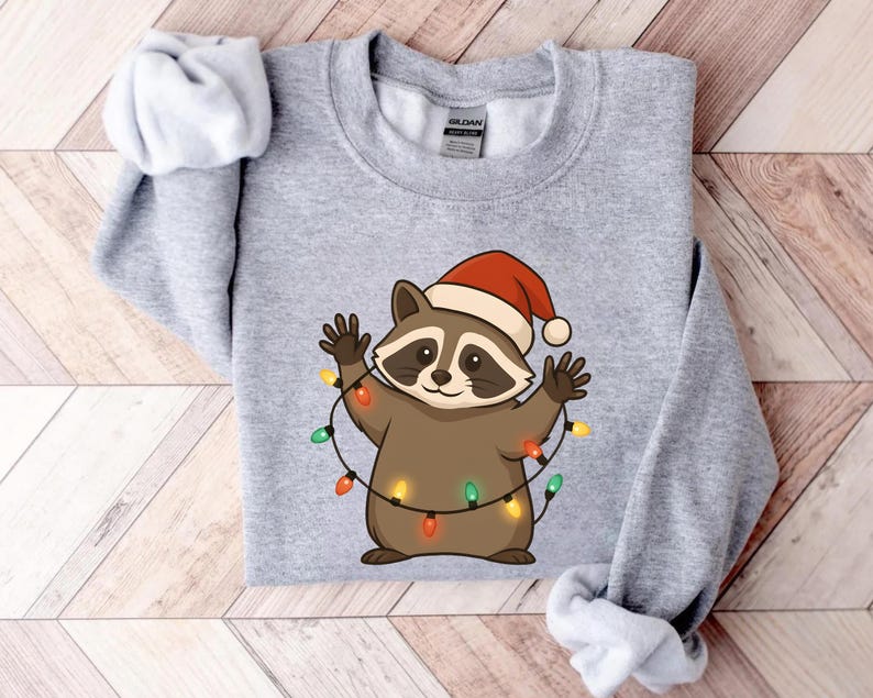Puede incluir: Sudadera gris claro con un mapache de dibujos animados con un gorro de Pap&aacute; Noel y una guirnalda de luces navide&ntilde;as de colores. El mapache tiene un cuerpo marr&oacute;n, una m&aacute;scara negra y est&aacute; sonriendo.