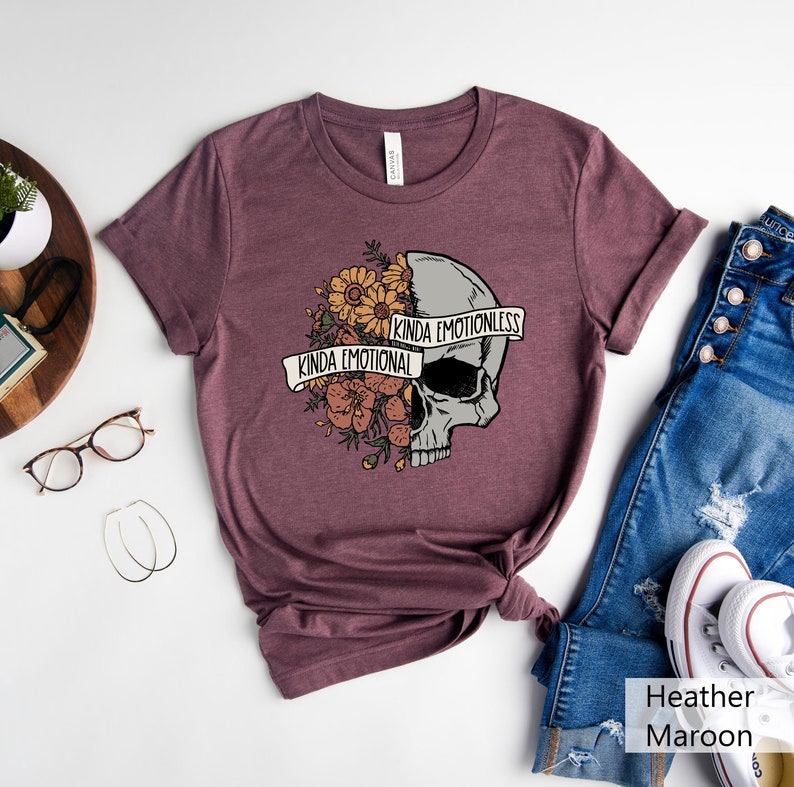 Pu&ograve; includere: Una t-shirt color marrone rossiccio con una grafica di un teschio. Il teschio &egrave; diviso a met&agrave;, con un lato ricoperto di fiori e il testo "Kinda Emotional" e l'altro lato con il testo "Kinda Emotionless".