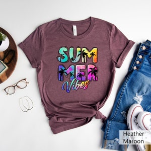 May include: A heather maroon t-shirt with the words "SUMMER VIBES" in a colorful, tropical design. The text features a gradient of blues, greens, yellows, and pinks, with black palm tree silhouettes. The shirt is folded and styled with jeans and white sneakers.