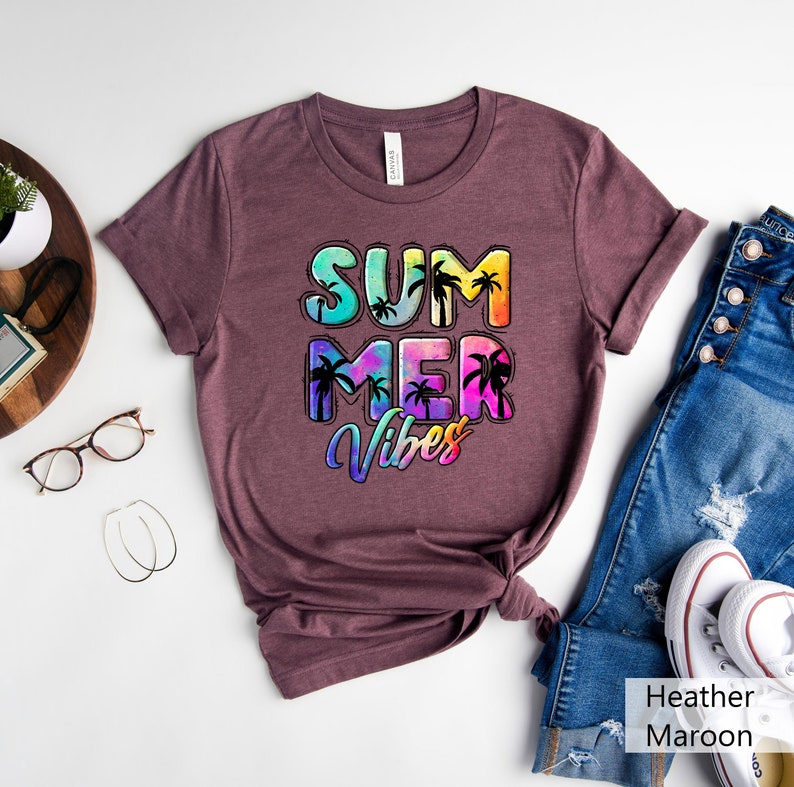 May include: A heather maroon t-shirt with the words "SUMMER VIBES" in a colorful, tropical design. The text features a gradient of blues, greens, yellows, and pinks, with black palm tree silhouettes. The shirt is folded and styled with jeans and white sneakers.
