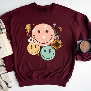 May include: A maroon sweatshirt with a graphic design of three smiley faces with stars and lightning bolts. The smiley faces are surrounded by flowers and leaves.