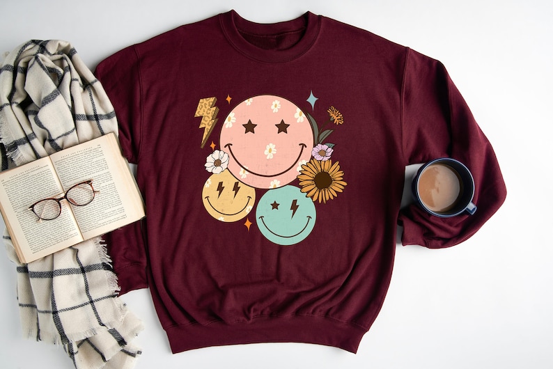May include: A maroon sweatshirt with a graphic design of three smiley faces with stars and lightning bolts. The smiley faces are surrounded by flowers and leaves.