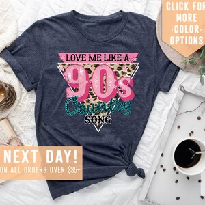 May include: A navy blue t-shirt featuring a pink and white graphic that reads "Love Me Like A 90's Country Song". The graphic is in a triangular shape with a leopard print background.