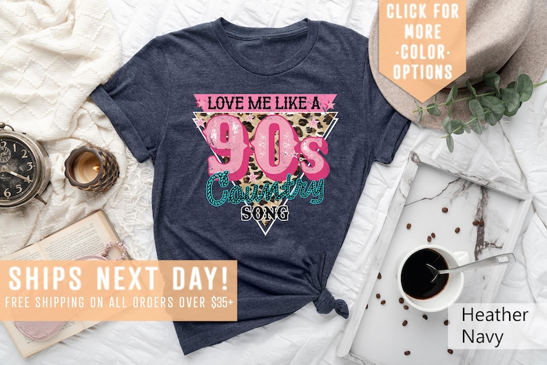 May include: A navy blue t-shirt featuring a pink and white graphic that reads "Love Me Like A 90's Country Song". The graphic is in a triangular shape with a leopard print background.