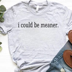 May include: A light gray t-shirt with the phrase "i could be meaner" printed in black. The shirt is paired with blue jeans and brown sandals. The shirt is made of a soft material.
