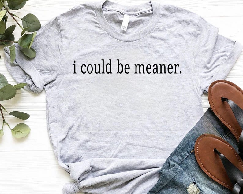May include: A light gray t-shirt with the phrase "i could be meaner" printed in black. The shirt is paired with blue jeans and brown sandals. The shirt is made of a soft material.