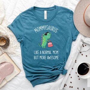 May include: Teal t-shirt with the text "MOMMYSAURUS" above a cartoon dinosaur wearing sunglasses and carrying a purse. The text "LIKE A NORMAL MOM BUT MORE AWESOME" is below the dinosaur. The shirt is displayed on a white surface.