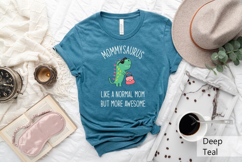 May include: Teal t-shirt with the text "MOMMYSAURUS" above a cartoon dinosaur wearing sunglasses and carrying a purse. The text "LIKE A NORMAL MOM BUT MORE AWESOME" is below the dinosaur. The shirt is displayed on a white surface.