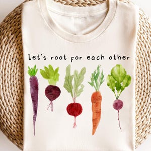 May include: Cream-colored t-shirt featuring a watercolor illustration of various vegetables, including carrots, beets, and radishes. The text "Let's root for each other" is printed above the vegetables. The shirt has a round neckline.