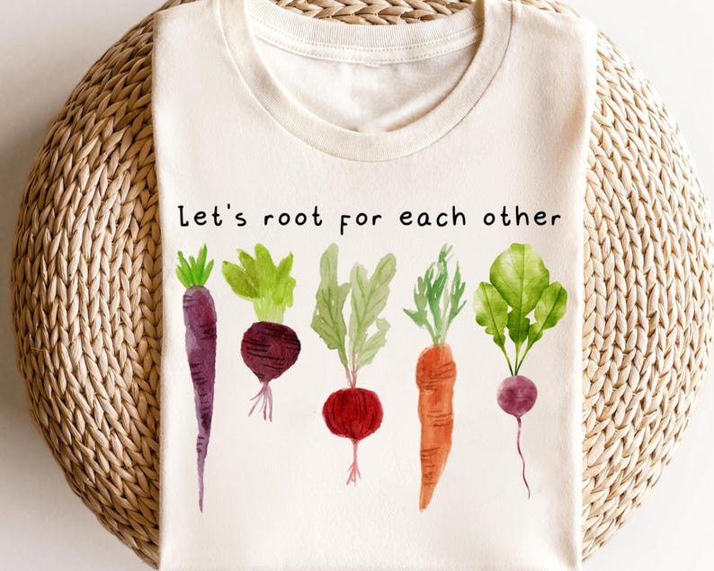 May include: Cream-colored t-shirt featuring a watercolor illustration of various vegetables, including carrots, beets, and radishes. The text "Let's root for each other" is printed above the vegetables. The shirt has a round neckline.