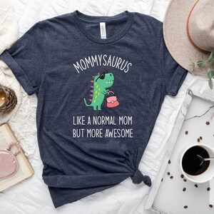 May include: Heather navy t-shirt with the text "MOMMYSAURUS" above a cartoon dinosaur wearing sunglasses and holding a purse. The text "LIKE A NORMAL MOM BUT MORE AWESOME" is below the dinosaur. The shirt is knotted at the bottom.
