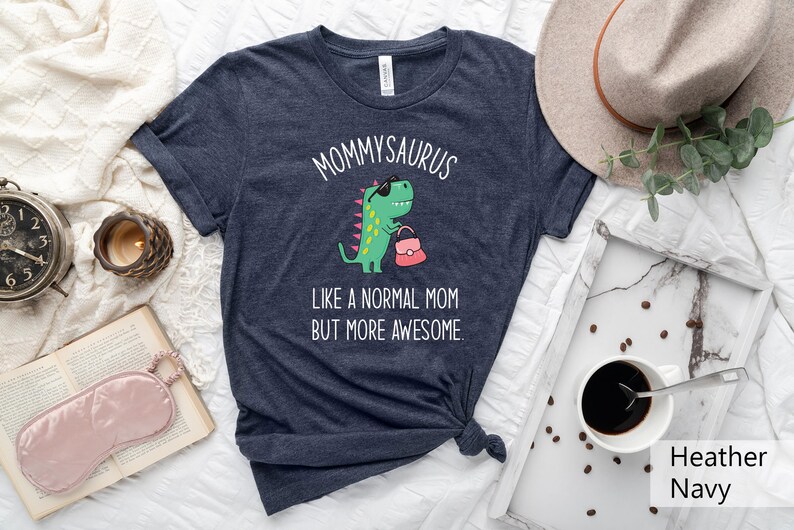 May include: Heather navy t-shirt with the text "MOMMYSAURUS" above a cartoon dinosaur wearing sunglasses and holding a purse. The text "LIKE A NORMAL MOM BUT MORE AWESOME" is below the dinosaur. The shirt is knotted at the bottom.