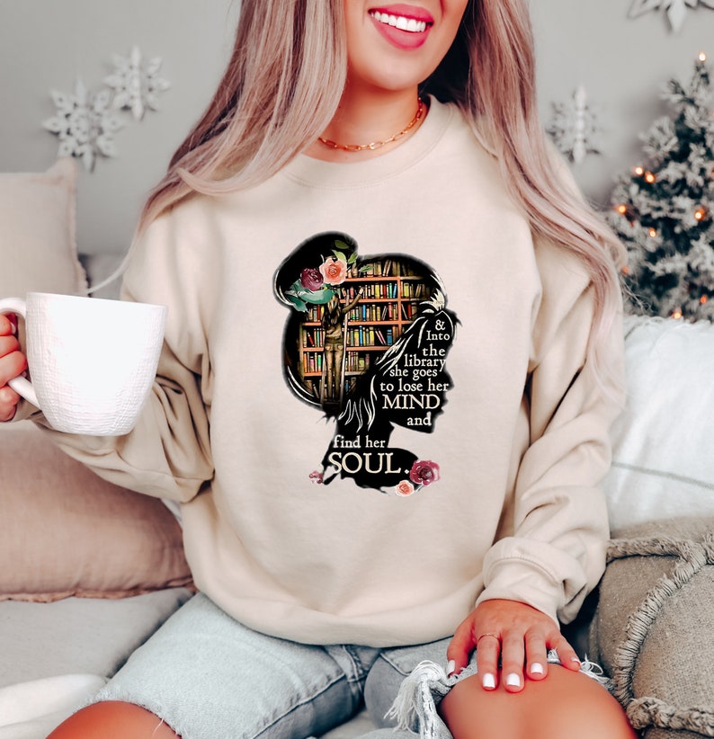 May include: A cream-colored sweatshirt with a graphic of a woman's silhouette with a bookshelf behind her. The text reads "& Into the library she goes to lose her MIND and find her SOUL."