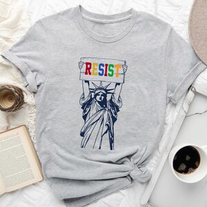 May include: A gray t-shirt with a graphic of the Statue of Liberty holding a sign that says "RESIST" in rainbow colors.