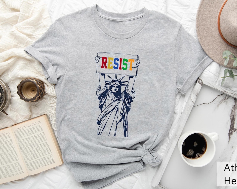 May include: A gray t-shirt with a graphic of the Statue of Liberty holding a sign that says "RESIST" in rainbow colors.