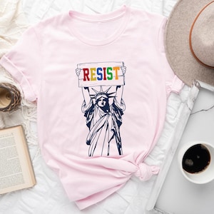 May include: Pink t-shirt with a graphic of the Statue of Liberty holding a sign that says "RESIST" in rainbow colors.