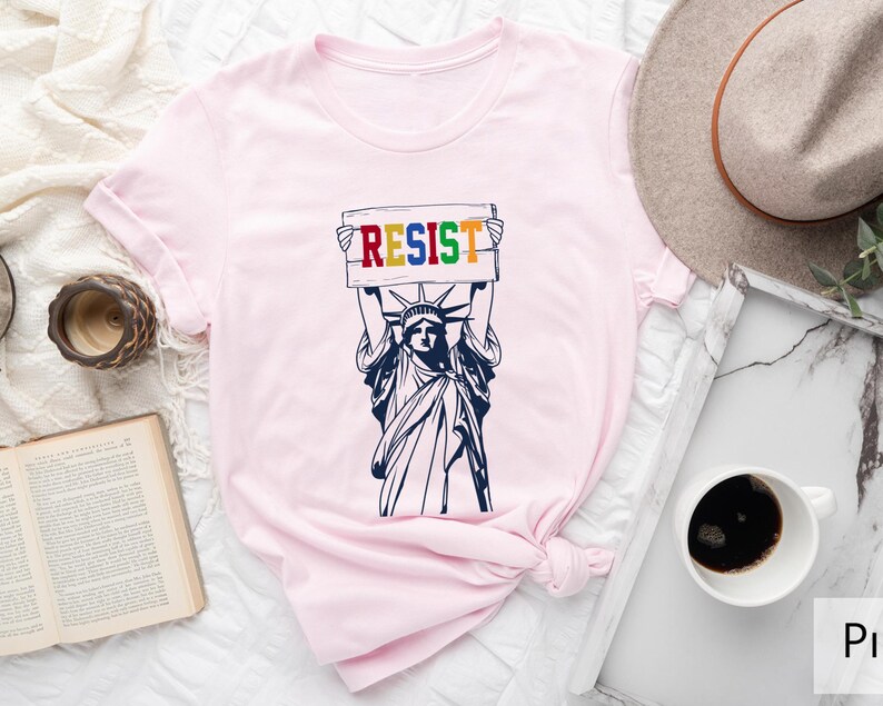 May include: Pink t-shirt with a graphic of the Statue of Liberty holding a sign that says "RESIST" in rainbow colors.