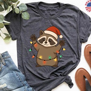 Puede incluir: Camiseta gris oscuro con un mapache de dibujos animados que lleva un gorro de Pap&aacute; Noel y sostiene una cadena de luces navide&ntilde;as de colores. El mapache es marr&oacute;n y canela con una m&aacute;scara negra. La camiseta se combina con vaqueros azules y sandalias marrones.