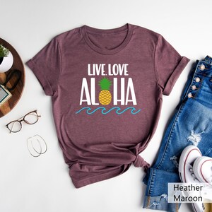 May include: Heather maroon t-shirt with the words "LIVE. LOVE. ALOHA" in white, a yellow pineapple with green leaves, and blue waves. The shirt is paired with blue jeans and white sneakers.