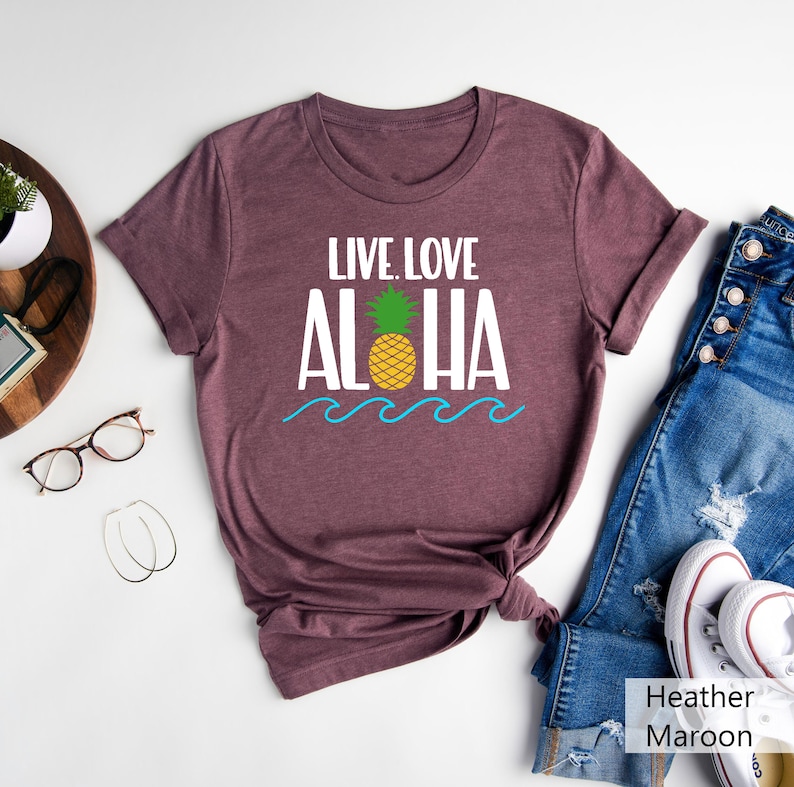 May include: Heather maroon t-shirt with the words "LIVE. LOVE. ALOHA" in white, a yellow pineapple with green leaves, and blue waves. The shirt is paired with blue jeans and white sneakers.