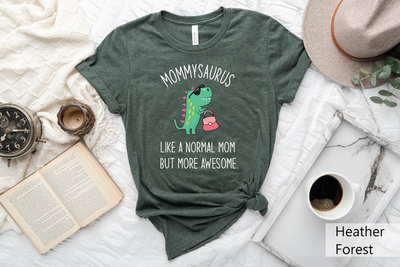 May include: Heather forest green t-shirt with the text "MOMMYSAURUS" above a cartoon dinosaur wearing sunglasses and carrying a pink purse. The text "LIKE A NORMAL MOM BUT MORE AWESOME" is below the dinosaur.