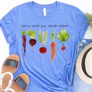 May include: A light blue t-shirt with the text "Let's root for each other" above a watercolor illustration of various root vegetables. The vegetables include a purple carrot, beet, radish, and carrot.