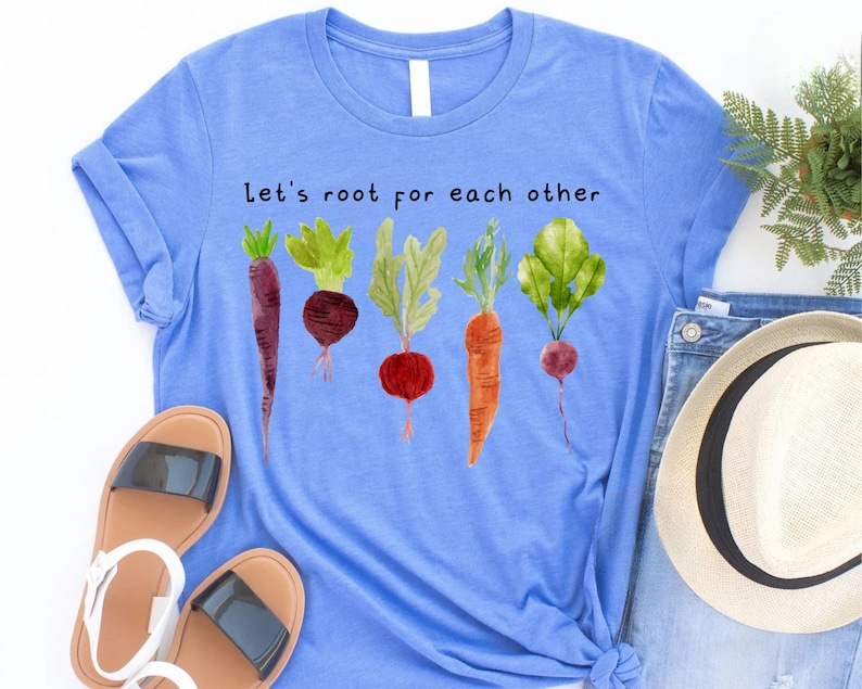 May include: A light blue t-shirt with the text "Let's root for each other" above a watercolor illustration of various root vegetables. The vegetables include a purple carrot, beet, radish, and carrot.