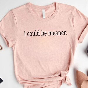 May include: A light pink t-shirt with the phrase "i could be meaner" printed in black. The shirt has short sleeves and a round neckline. The image also shows a pair of glasses, a scarf, and a pair of shoes.
